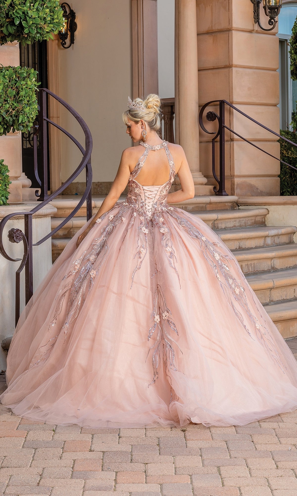 A woman in the Sleeveless Quinceanera Dress 1677, featuring pink floral embroidery and a corset back, stands on a stone path before a staircase, facing away from the camera.